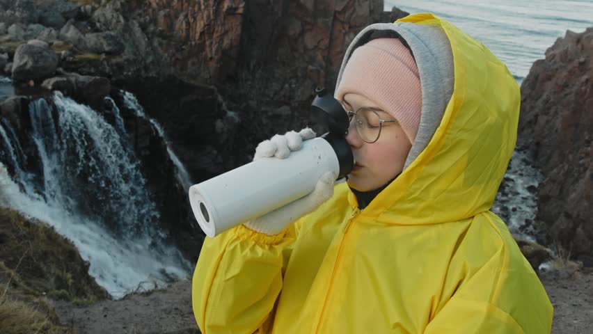 A tourist stands beside a mighty waterfall, but the chill in the air prompts her to sip on a warm beverage from her portable mug. Portrait.