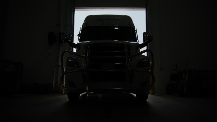 Truck drives backward from the auto repair shop with emergency lights on, Indoors wide shot. 