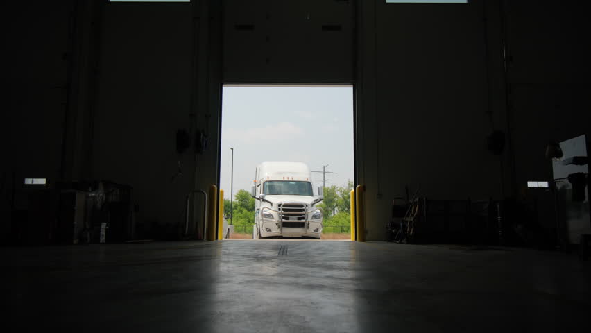Truck drives forward into auto repair shop with emergency lights on, Indoors wide shot. 