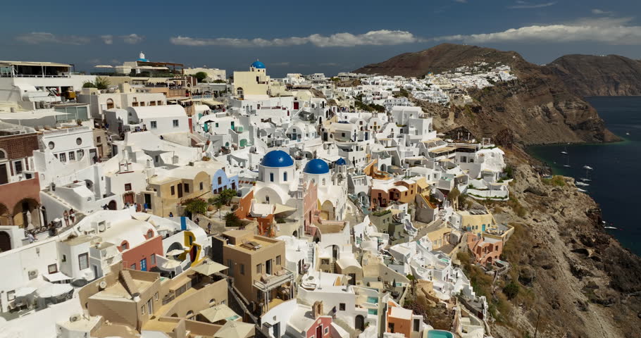 Flying backwards above famous view point on Santorini Island in Greece. People watching blue dome churches and white washed buildings