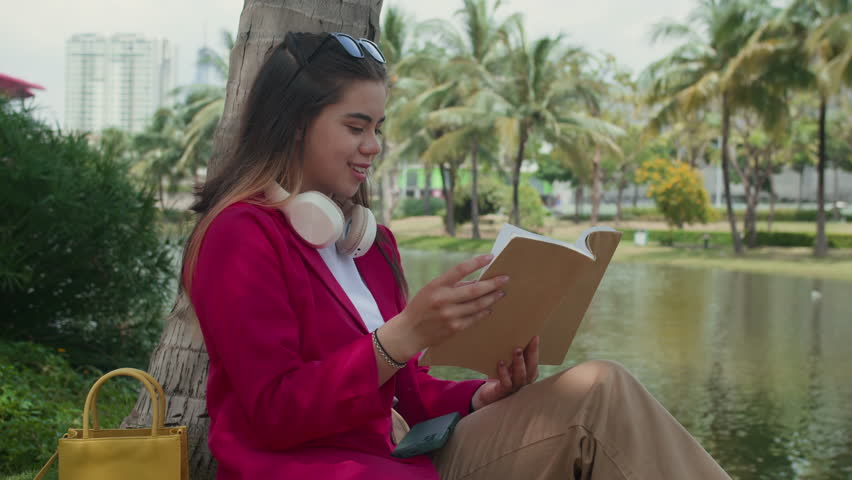 Medium long shot of female multicultural zoomer having rest in park while sitting by tree and reading book
