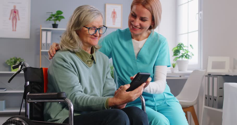Caregiver helping an elderly patient in a wheelchair at a nursing home use a mobile phone and internet. Providing assistance with online technology to foster connection and engagement.