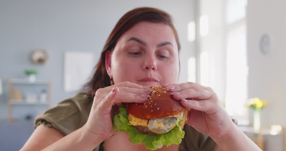 Obese hungry happy woman with excessive fat eating unhealthy fast food, tasty hamburger, juicy burger, cheeseburger, chubby overweight plus size big fat fit lady with good appetite, excess calories 