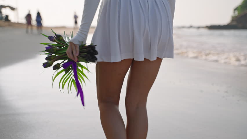 Female feet going in sea surf waves. Pretty model walking and smelling bouquet of purple flowers along coast of tropical island. Romantic woman strolling on the ocean shore. Close-up, back view.
