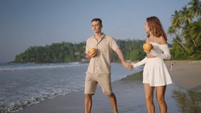 Happy couple walk, drink coconut water on ocean beach. Young woman, man hold hands, kiss on stroll. Romantic newlyweds clink cocktails, enjoy honeymoon vacation at tropical sea shore on sunrise. - Powered by Shutterstock - Get 15% off with code: PIKWIZARD15