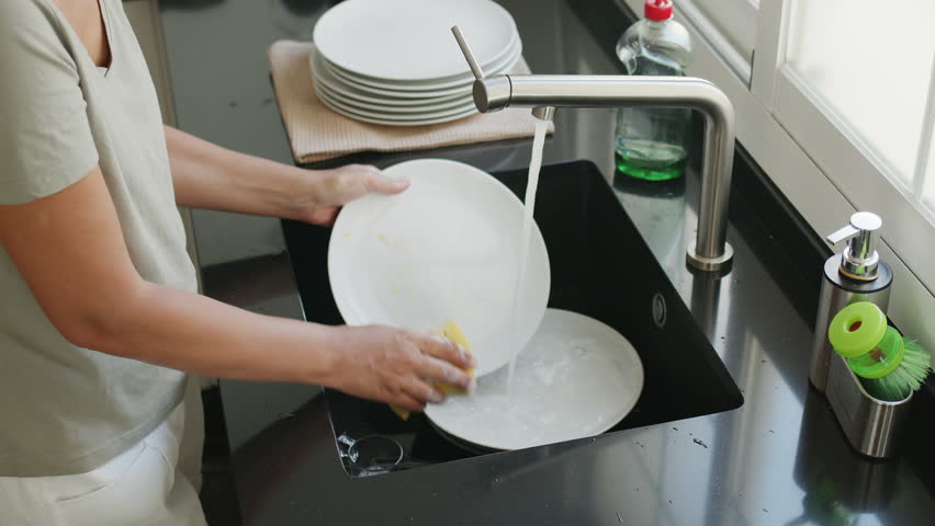 Woman washing and rinsing dishes by hand under a stream of water in the kitchen sink. Washing dishes process. Slow motion video. 