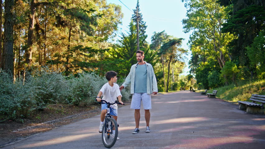 Caring dad teaching kid to ride bicycle at greenery recreational zone. Attentive boy listening man trying to cycling at sunshine forest. Positive son learning new sport with father at green park 
