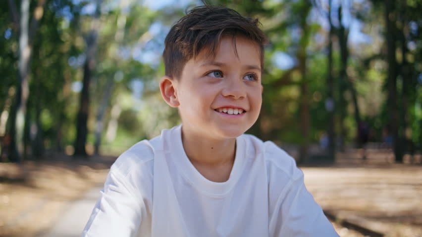 Cheerful kid looking camera at sunshine forest portrait. Happy smiling child feeling positive having fun at sunny greenery park closeup. Brunette small boy enjoying holidays spending weekend outdoors