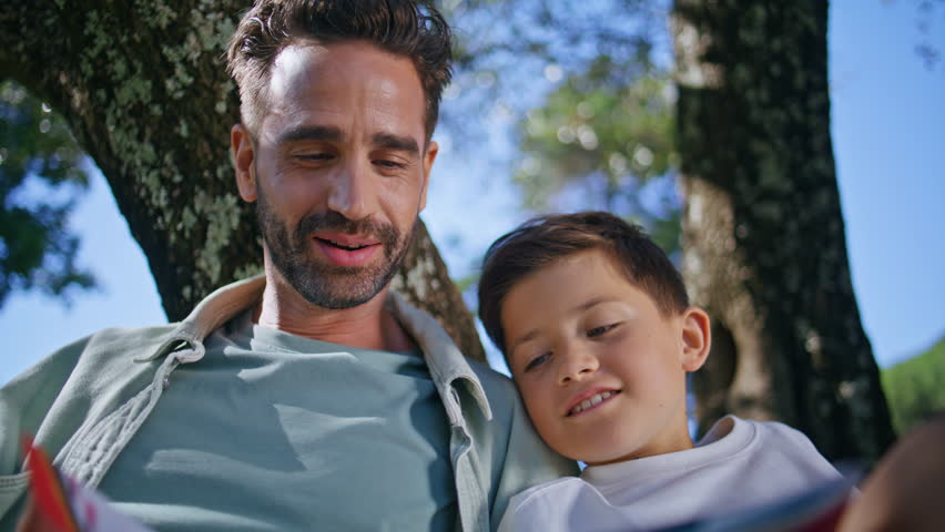 Father son flipping pages sitting under tree at sunny park closeup. Happy family reading book outdoors on summer picnic. Cheerful parent child bonding together on green nature place. Childhood concept