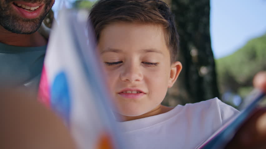 Bonding family reading fairytale spending free time at fresh air closeup. Positive parent son enjoying carefree lifestyle sitting under tree. Small kid watching pictures in book relaxing at forest