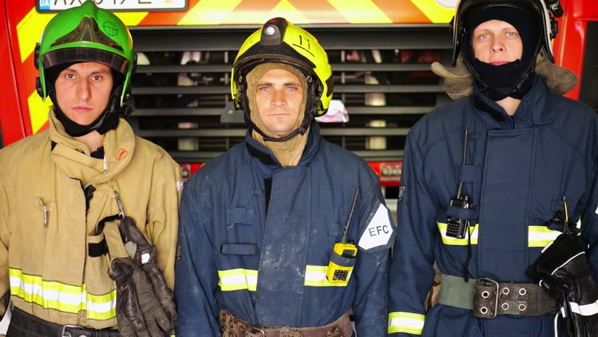 Portrait of male firefighters in helmets and protective uniforms standing near a big red car. Young confident firemen in full equipment posing against the background of a fire engine at station