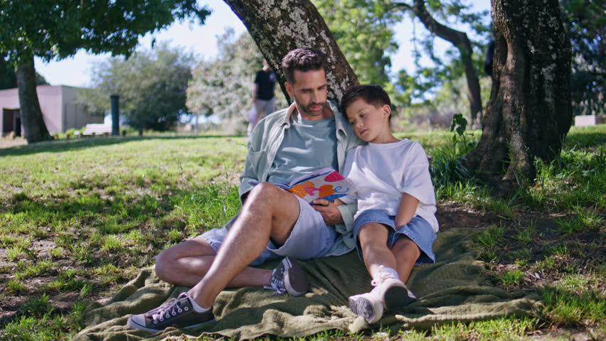 Dad kid watching book sitting green grass at forest. Adorable little boy expressing happiness spending weekend with father resting under big tree. Cute loving family of parent son reading fairytale 