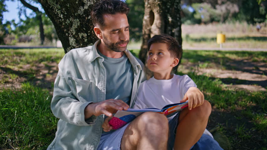 Cute family reading book sitting green grass at forest closeup. Adorable little kid boy having fun expressing happiness spending weekend with father under big tree. Loving parent son relationships 