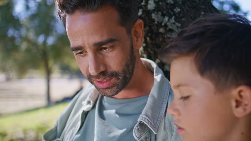 Bearded father talking kid sitting under tree at garden closeup. Caring daddy little child enjoying communication at summer nature. Small boy listening man at cozy park. Parenthood childhood concept