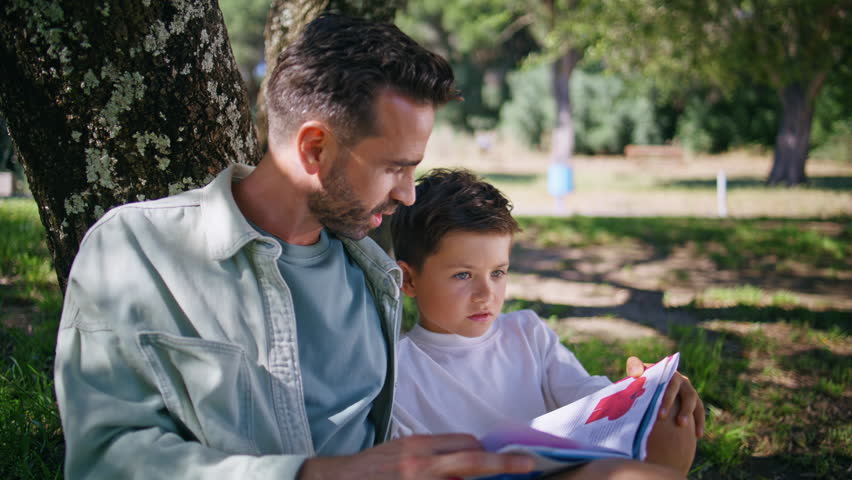 Daddy child reading fairytale gathering in forest nature closeup. Bearded parent man and kid boy resting on green grass enjoying book. Caring father and son bonding under tree talking together at park