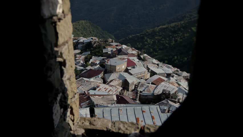 Warm sunlight filtering through cracked stone window, revealing stone-roofed village nestled amid lush green mountainside, depicting peaceful rural tranquility
