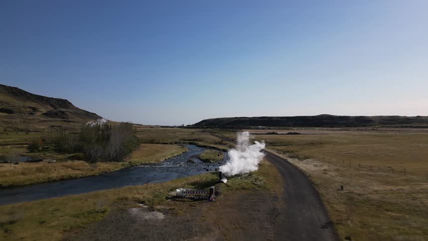 Iceland views. A river flows through a grassy field with a few trees in the background. The sky is clear and blue, and the sun is shining brightly