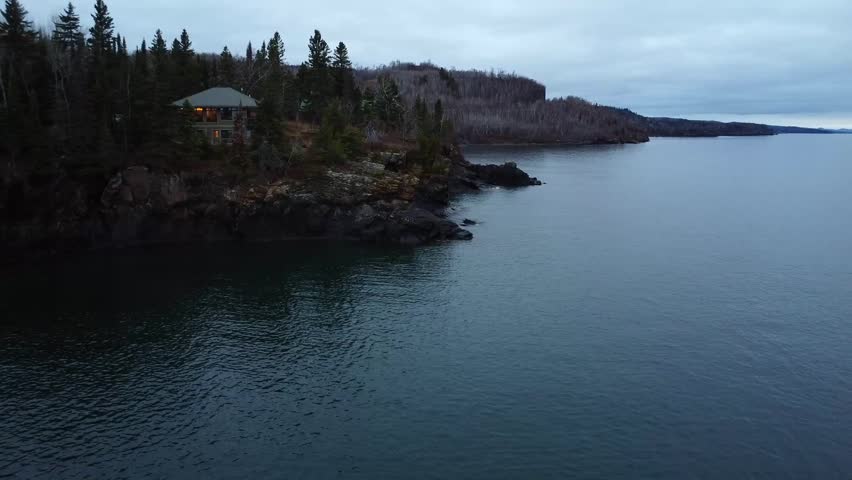 Video shot of serene misty landscape, a house on rocky cliffs and a forest with leafless trees in the fall season. A calm lake in Minnesota.