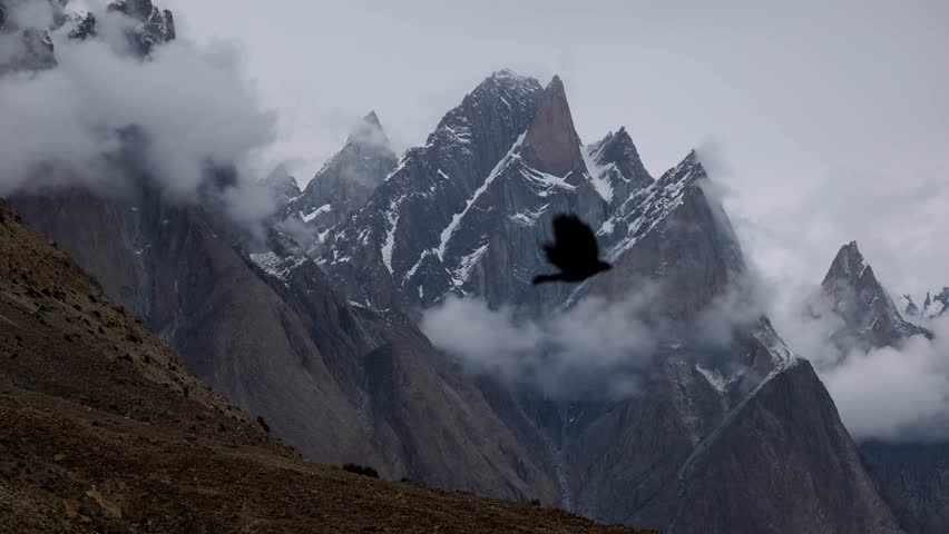 Pointy triangular mountains in Pakistan shrouded in low-hanging clouds with a few birds flying by. Captured while trekking to K2 Base Camp.