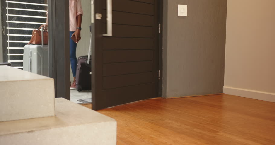 Carrying suitcase, african american woman entering home with handbag, wearing casual clothes. Travel, luggage, arrival, domestic, unpacking, journey