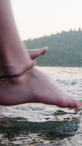 Vertical close-up view of legs of two unrecognisable young people wearing ankle bracelets splashing water while sitting on lake pier