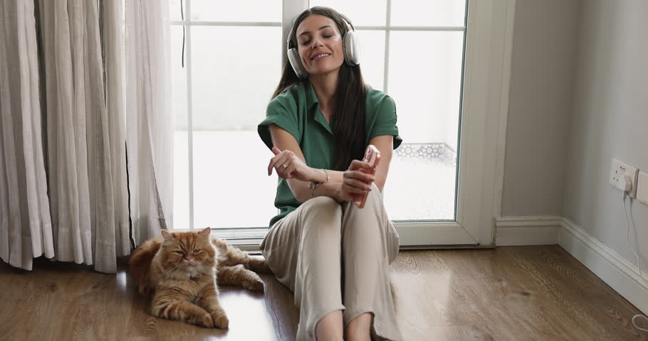 Serene woman listening favourite music through modern ambient wireless headphones seated on warm floor with cute ginger cat nearby, spend carefree weekend rest together at modern home. Hobby, leisure