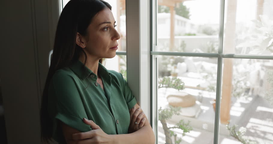 Close up woman gazes out window with sad and reflective expression, spend time alone at modern home, thinks on personal problems, break up, missing, waits for someone. Loneliness, solitude, concerns