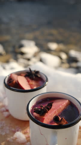Vertical footage of two mugs with hot mulled wine or gluhwein with spices and orange slices on wooden board near winter river at sunny day. Traditional drink on winter holiday