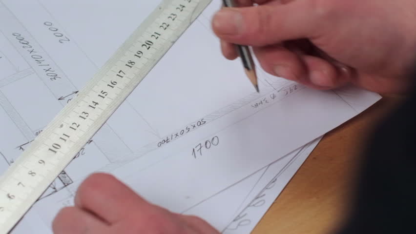 Close-up of hands working on technical drawings. A metal ruler is being used to measure or draw precise lines on a sheet of paper containing architectural or construction sketches