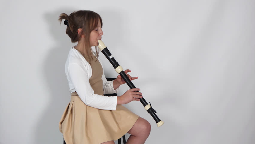 Girl playing, practicing the flute, playing a melody on a musical instrument, wooden pipe, sitting on black chair on white background. Out-of-school children activities, lessons