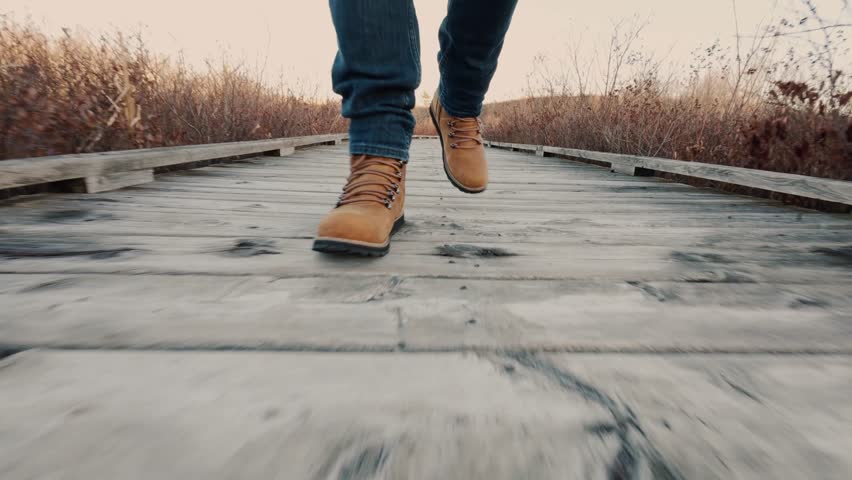 Man In Hiking Boots Walking Across A Wooden Suspension Bridge Over A Stunning Mountain Lake. An Epic Hiking Journey That Takes You Through Untamed Wilderness.