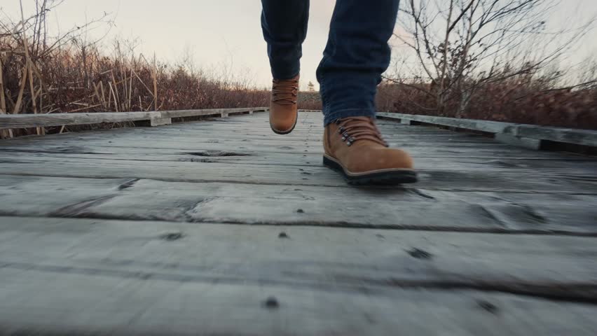 A Man In Hiking Boots Walks Across A Wooden Suspension Bridge Over A Wild Mountain Lake. Adventure Provides A Close-Up View Of The Thrills Of Hiking And Outdoor Exploration.