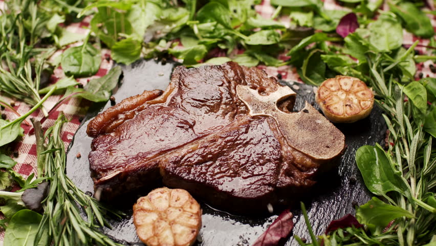 Grilled T-Bone Beef Steak Being Salted with Greenery Around