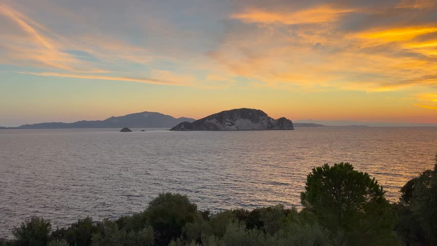 Breathtaking sunset over Marathonisi Island in Zakynthos, Greece, with vibrant clouds, water reflections, and lush foreground greenery. PAN RIGHT