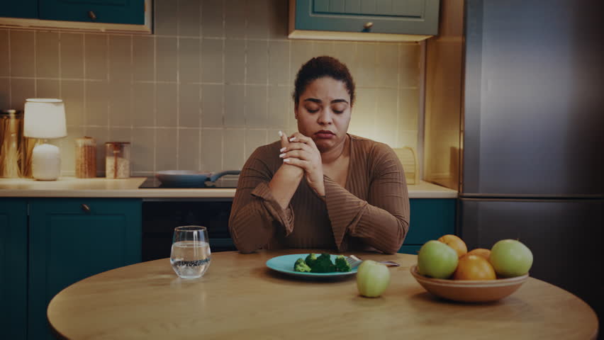 Irritated African American woman looks at broccoli on plate trying to eat meal. Overweight lady reflects unwillingness to follow healthy diet according to doctor advice