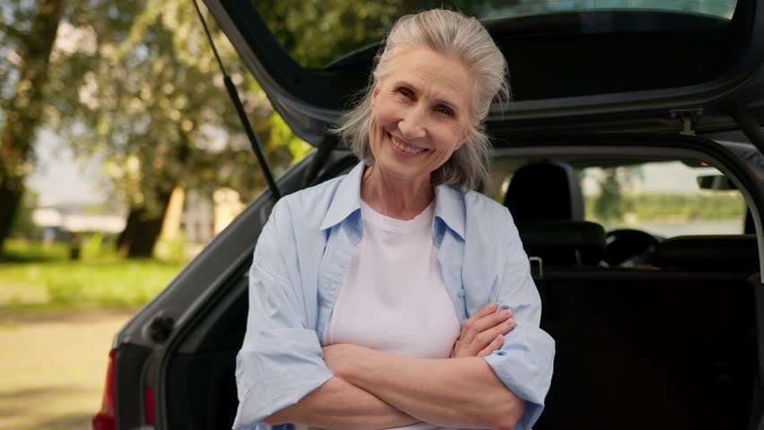 Joyful confident successful Caucasian mature elderly grandmother woman female senior car auto driver lady sitting automobile vehicle trunk looking at camera crossing hands crossed arms outside nature