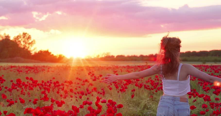 Beautiful young girl running in flower poppy field at sunset, slow motion. Landscape with lots of red flowers in meadow, natural idyllic spring sunset
