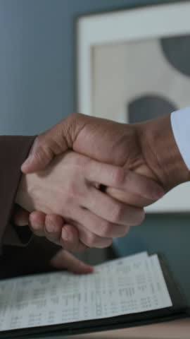 Vertical close-up shot of hands of unrecognizable woman holding financial report and shaking hands with business consultant during meeting in office