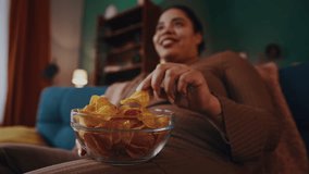 African American woman eats chips watching TV show on sofa in evening. Plus size female suffers from unhealthy habit experiencing eating disorder - Powered by Shutterstock - Get 15% off with code: PIKWIZARD15