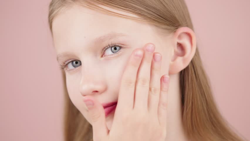 Portrait of a beautiful cute young girl taking care of her face skin. Blonde Caucasian girl with blue eyes isolated on pink background. Advertising concept of skin care for teenagers. Acne treatment.
