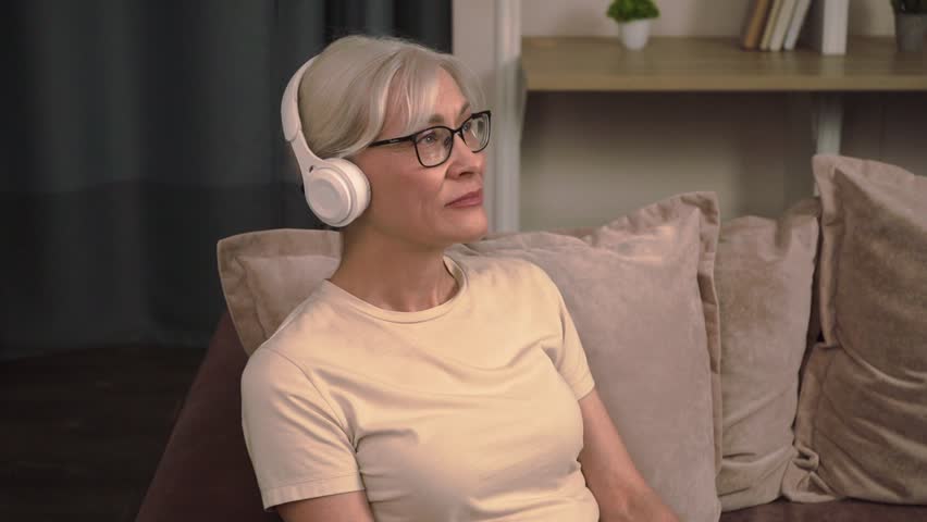 Old woman wearing headphones, sitting on a couch with a tablet, using technology for video calls, staying connected loved ones 