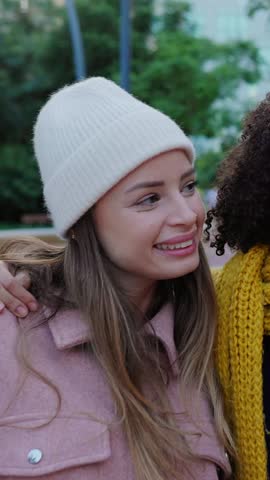 Vertical HD video of three happy young teenage girls having fun hugging each other at city street, bundled up in cozy warm clothes. Youth and female friendship concept
