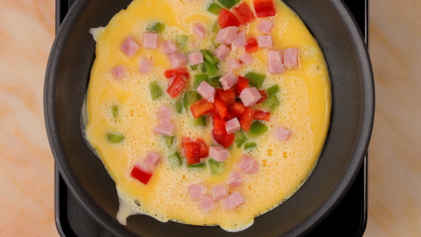 Eggs are poured into frying pan and cooked, hand adds peppers, ham, cheese and