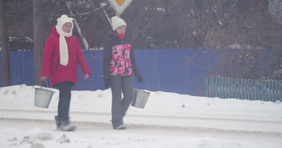 Two girls dressed in warm winter clothing carry metal buckets filled with water from a water tower, walking through snowy terrain to prepare baths, showcasing traditional rural practices.