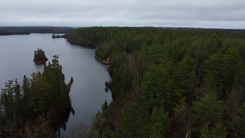 Peaceful aerial view of a forest-lined lake in the Boundary Waters Canoe Area Wilderness, featuring calm waters and dense evergreen trees under an overcast sky