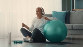 A cheerful elderly lady engages in a workout session at home, using a stability ball for exercise. She wears headphones and enjoys her fitness routine in a bright, cozy space. - Powered by Shutterstock - Get 15% off with code: PIKWIZARD15