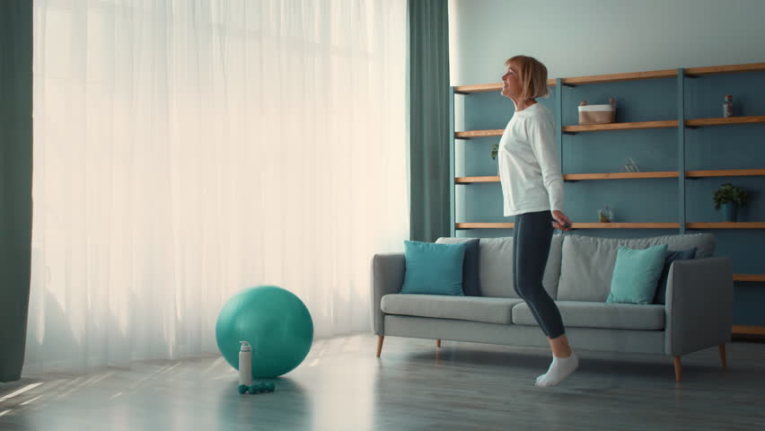 An elderly woman exercises indoors in a bright room. She performs physical activity with a blue exercise ball nearby, showcasing her dedication to fitness and well-being.