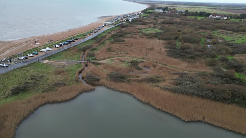 An aerial view of a coastal nature reserve. A winding path leads through a diverse landscape of tall reeds, lush greenery, and a tranquil pond.