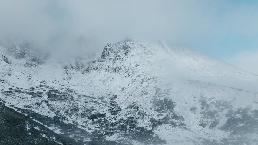 Aerial shot of a snow-covered mountain (Lomnicky stit, Slovakia) ridge partially veiled by clouds, creating a serene and majestic alpine atmosphere. (4K)