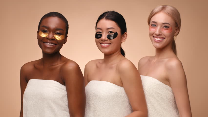 Three young women stand close together wearing towels and eye patches while smiling. They are engaging in a skincare routine, sharing a joyful moment in a warm setting.